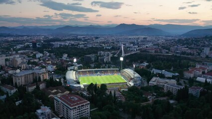 Cityscape featuring a stadium with green grass soccer field (football pitch) illuminated by floodlights at night, in the dark city of Podgorica, Montenegro, under colorful sunset sky in the evening. - Powered by Adobe