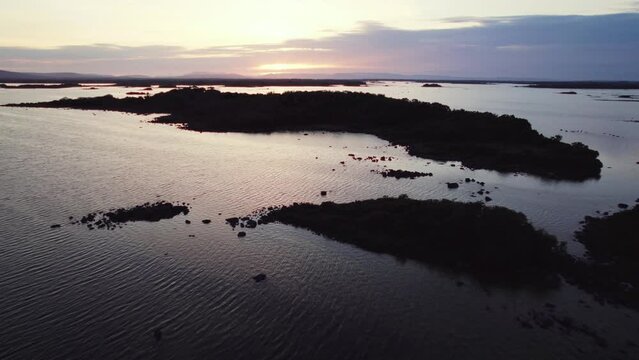 Beautiful aerial flying over gigantic irish lough corrib with many small island silhouettes in the water. Flying forward, tilt down and up again during blue hour golden evening sun reflecting in waves