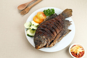 Gurame Bakar Madu, Grilled Gourami with honey and soy sauce. Served with sambal (chilli sauce), cucumber and tomatoes in white plate.   
