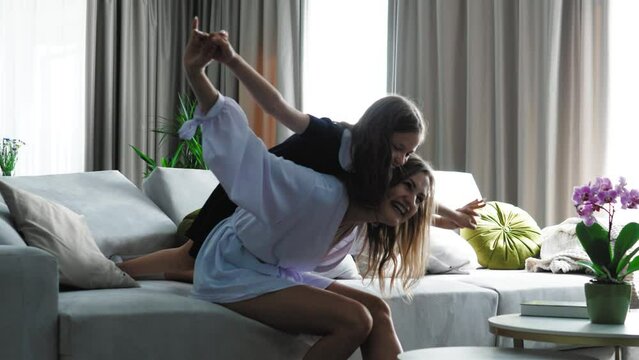 Happy family loving young mum playing piggybacking cute little funny kid daughter sit on couch at home, cheerful mother carrying small child girl on back bonding pretending flying having fun together