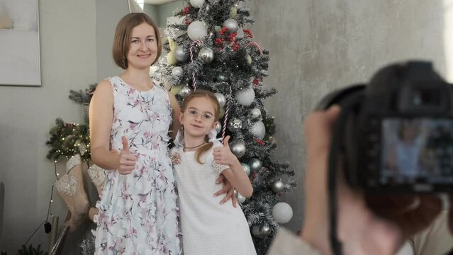Young woman with little daughter posing over Christmas tree while her husband taking photos on digital camera at home