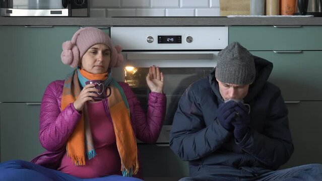 Young man and woman in outwear trying to warm up by oven at home, drinking hot drinks while sitting on the floor in the kitchen. Energy and gas crisis, heating problems concept