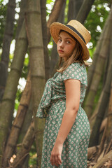 Vintage style portrait of a Young Girl with a green dress and a straw hat standing up in  a bamboo forest
