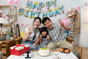 happy asian small family looking at camera with victory hand gestures at the baby girl's second birthday celebration party at home with pink balloon and garland