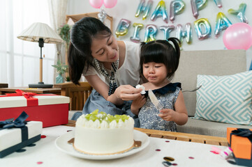 smiling asian mother wiping baby girl's face with a tissue as she is playing with a cake server at the birthday party at home