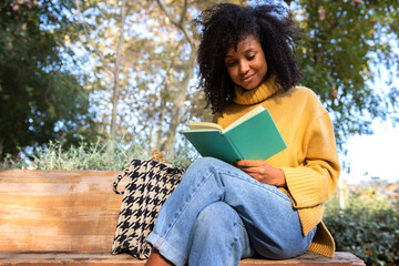 Happy young African American woman sit on a park bench reading a book. Copy space.