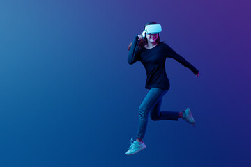 Naklejka na ściany i meble Young Asian woman wearing VR headset with experience playing video game and jumping levitating in the air on futuristic purple cyberpunk neon light background. Metaverse technology concept.