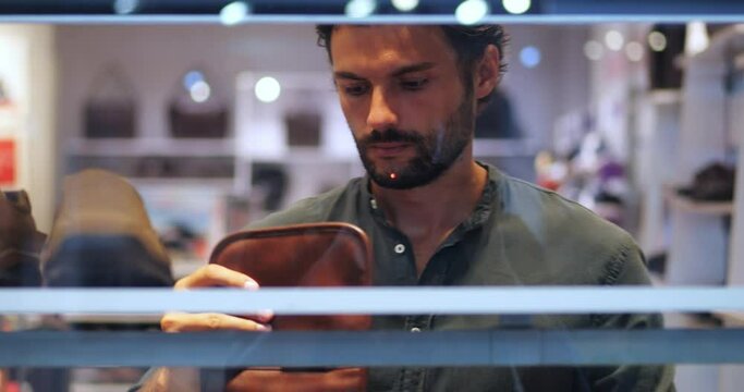 View through the shelves of clothes and accessories of a fashionable men's clothing store, of a handsome young dark-haired attractive man examining leather purses and making purchases in the mall