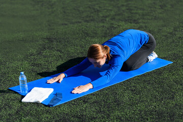 Women and sport. Girl in sportswear - blue shirt and black leggings does exercises: stretching out on the mat on the grass at the stadium on a sunny day. Middle aged sportswoman 