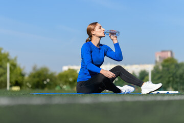 Women and sport. Girl in sportswear - blue shirt and black leggings resting on a mat on the grass and drinks water from a plastic bottle at the stadium outdoor on a sunny day. Middle aged sportswoman 