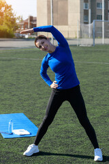Women and sport. Girl in sportswear does exercises: bends and stretches on the grass at the stadium on a sunny day. Middle aged sportswoman dressed in sportsclothes exercising outdoors