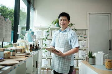 Portrait of an Asian male shopkeeper smiling and looking at camera, arranges natural products at refill store, zero-waste grocery, and plastic-free, eco environment-friendly, sustainable lifestyles.