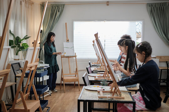 A Female Asian Teacher Teaches And Demonstrates To The Children On Acrylic Color Picture Painting On Canvas In Art Classroom, Creatively Learning With Skill At The Elementary School Studio Education.