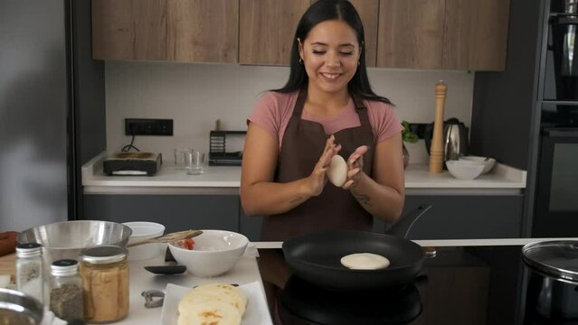 Young Venezuelan woman moulding the dough into a arepa and cooking it in a pan at kitchen.