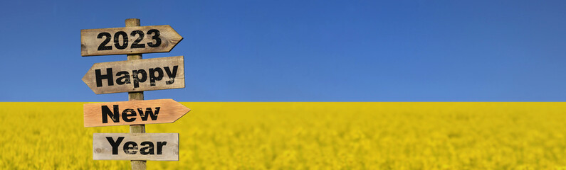 2023 happy new year written on a direction sign in front of field of yellow colza rapeseed blooming under blue sky  colors of ukrainian flag