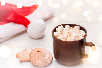 Fototapeta na wymiar mug with marshmallows, santa hat and cookies on the table close-up
