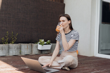 Beautiful girl sit on floor of terrace and take sips of fresh orange juice and look away, side...