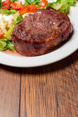 Cooked thin juicy beef steak on a white plate with green salad on a wooden table. Premium high quality meat dinner. Soft and tender meat cut prepared to highest standard. Close up.