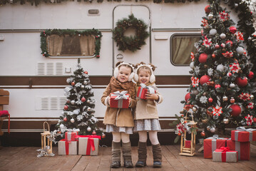 two little twin girls dressed in stylish fur coats play, hug and enjoy near the trailer in the...