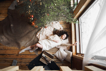 Fototapeta na wymiar Young stylish girl lies on skin on floor in cozy wooden country house,chalet in winter forest. Celebrating new year eve. Gifts,family holiday. Romantic weekend Christmas tree with gold red xmas balls