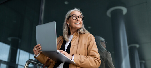 Business senior woman working on a laptop online against the backdrop of an office building