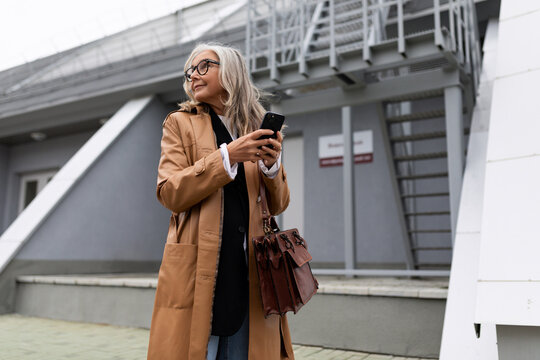 Mature Gray-haired Stylish Business Woman Rushing To A Meeting With A Mobile Phone In Her Hands