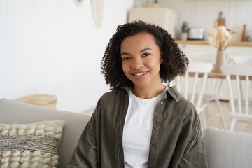 Smiling young mixed race girl with afro hairstyle sits on sofa in cozy living room at home