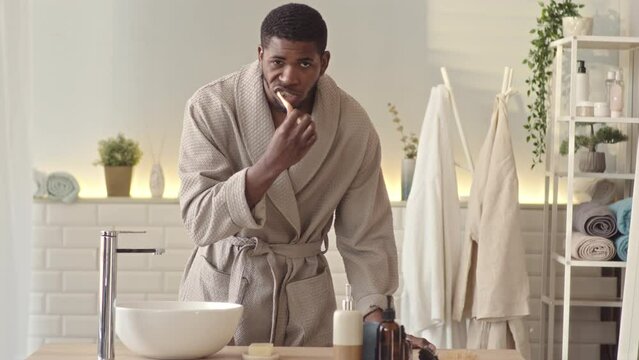 Medium slowmo of brutal young handsome African American man in bathrobe looking at his smartphone while brushing teeth in bathroom