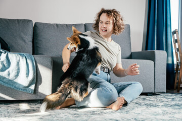 Curly haired glad, cheerful barefoot woman with dog corgi. Obedience training, feeding of pet, sitting, relaxing and playing on floor in comfortable living room near sofa. Weekend leisure activity