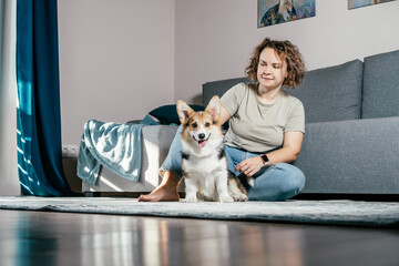 Curly haired glad, cheerful barefoot woman with dog corgi sitting, relaxing and playing on floor in comfortable living room, near sofa. Active and healthy animal, healthcare. Weekend leisure activity