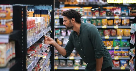 Thoughtful bearded attractive young man is shopping in the grocery department of a supermarket. Calculating expensive prices, looking aside on shelf with food, standing with a trolley in a supermarket - Powered by Adobe