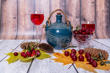 Autumn still life. On wooden table are earthenware jug, glasses of red wine, vase with ripe cherries and yellow maple leaves. Festive mood.