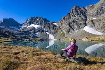 Fototapeta na wymiar The girl sits against the backdrop of mountain peaks and a blue lake. Beautiful mountain landscape for vacation, travel and healthy lifestyle