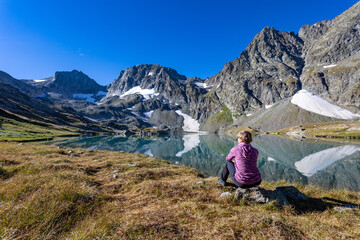 Naklejka na ściany i meble The girl sits against the backdrop of mountain peaks and a blue lake. Beautiful mountain landscape for vacation, travel and healthy lifestyle