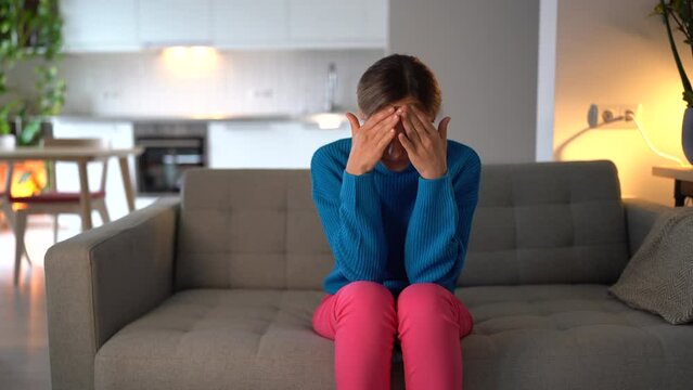 Unhappy Caucasian woman clutching head and touching face because of stress and depression sits on sofa of living room of apartment. Casual girl worry about high bills or impending financial problems