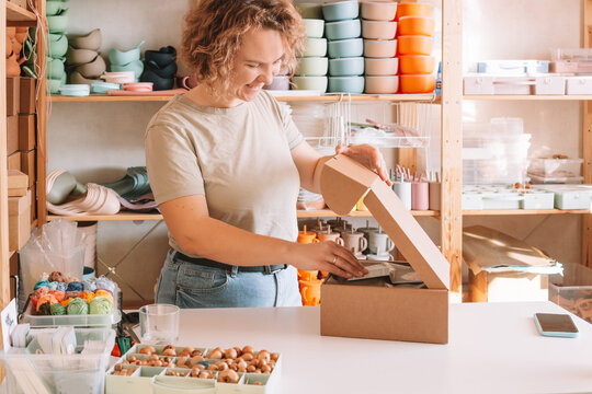 Smiling blond curly woman packing and wrapping shipment with postcards, silicone baby dishes in cardboard delivery box. Colorful skein of thread in container on desk. Workshop store, small business