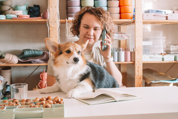 Smiling concentrated businesswoman talking smartphone, holding cute calm dog corgi in workshop. Set of silicone baby dishes near wall on shelves. Home office, taking orders in comfortable atmosphere