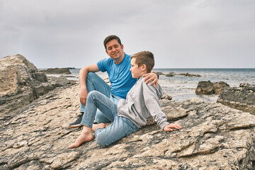 Fototapeta na wymiar Happy family, father and son bonding, sitting on stone by the sea looking at view enjoying summer vacation. Togetherness Friendly concept
