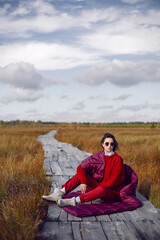 Fototapeta na wymiar portrait of a fashionable beautiful brunette woman sit on an ecological path in the marshes in autumn in a red sweater, hat and an unusual scarf. in Yelnya in Belarus.