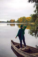 woman in a green sweater, hat and sunglasses stands on an old wooden boat by the lake in autumn in...
