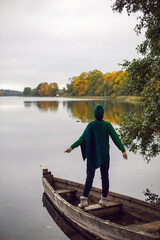 Fototapeta na wymiar woman in a green sweater, hat and sunglasses stands on an old wooden boat by the lake in autumn in the village. Vertical.