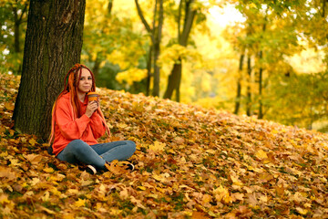 Young woman with dreadlocks drinking coffee from cup, sitting near tree in golden autumn. Vivid female with colourful hairstyle enjoying hot drink in nature.