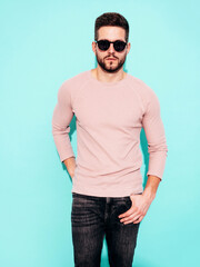 Fototapeta premium Portrait of handsome model. Sexy stylish man dressed in pink sweater and jeans. Fashion hipster male posing near blue wall in studio. In sunglasses. Isolated