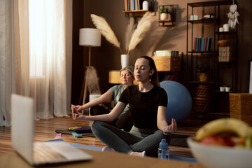 Yoga time in modern room young girl sitting on yoga mat in lotos pose closing eyes relaxing in front of laptop watching video trainig online.