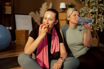 Delighted young girl biting ripe apple after workout with elderly mother. Middl-aged woman drinks water. Delighted women sitting on yoga mat relaxing indoors.