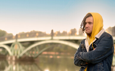 Portrait of young positive man with dreadlocks in hood, posing in city park on background of blurred bridge in cloudy weather