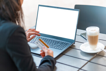 Young brunette business woman dressed in black suit spending her lunch time at street cafe terrace,drinking coffee.buy by credit card on her laptop.Blank white screen. Mockup,copy space for your text.