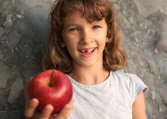 female child holds out an apple and smiles, no front tooth, gray stone background