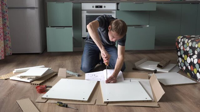 Young man assembling furniture for new apartment on the floor of kitchen, screwing screw into hole board