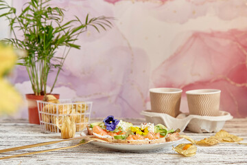 Fototapeta na wymiar Aesthetic lunch time - appetizer toasts with smoked salmon, avocado, arugula, cucumber and edible flowers. Cardboard cups with coffee. Cozy lagom home